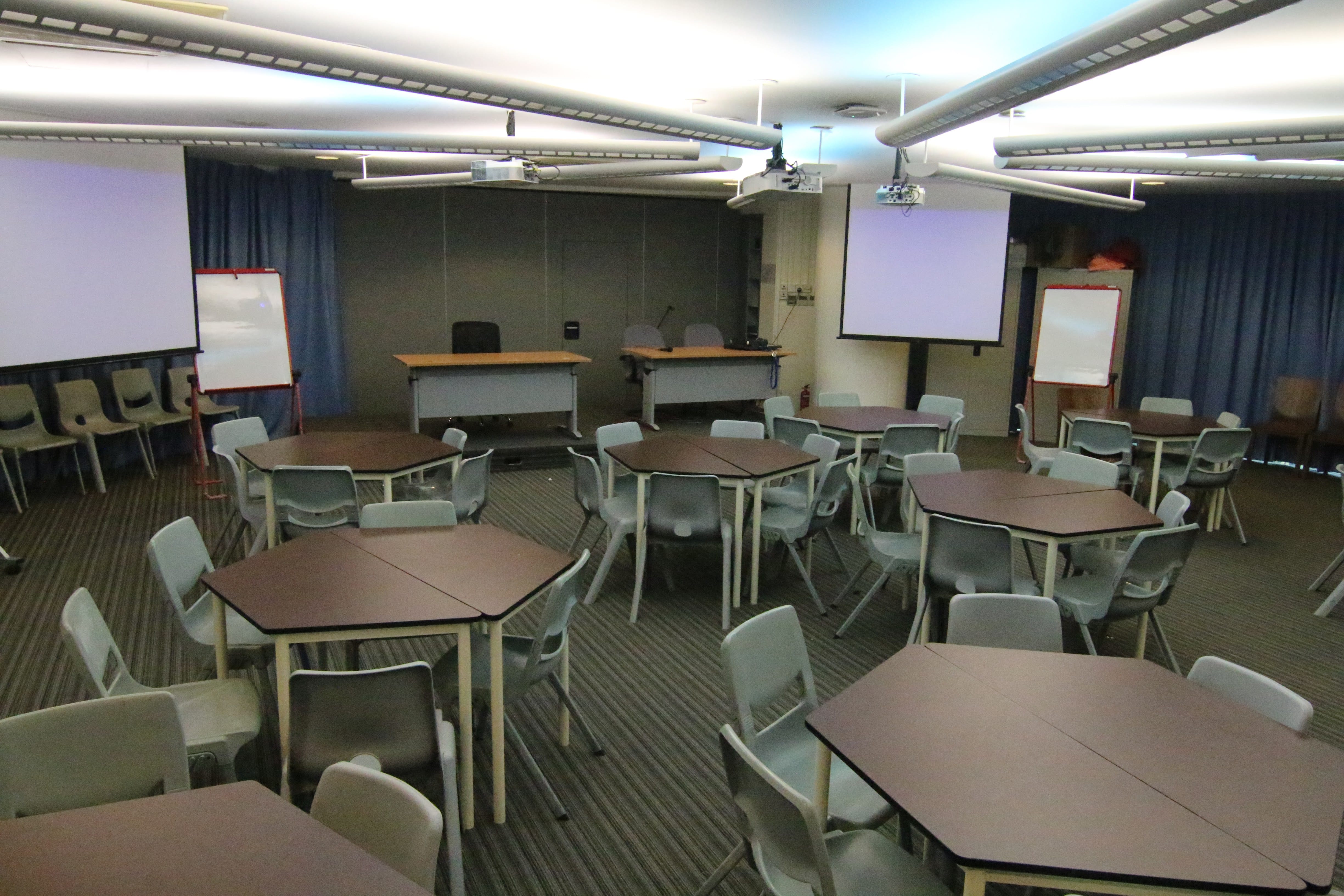 Empty classroom with hexagonal tables, gray chairs, a projector screen, and blue curtains along the wall.